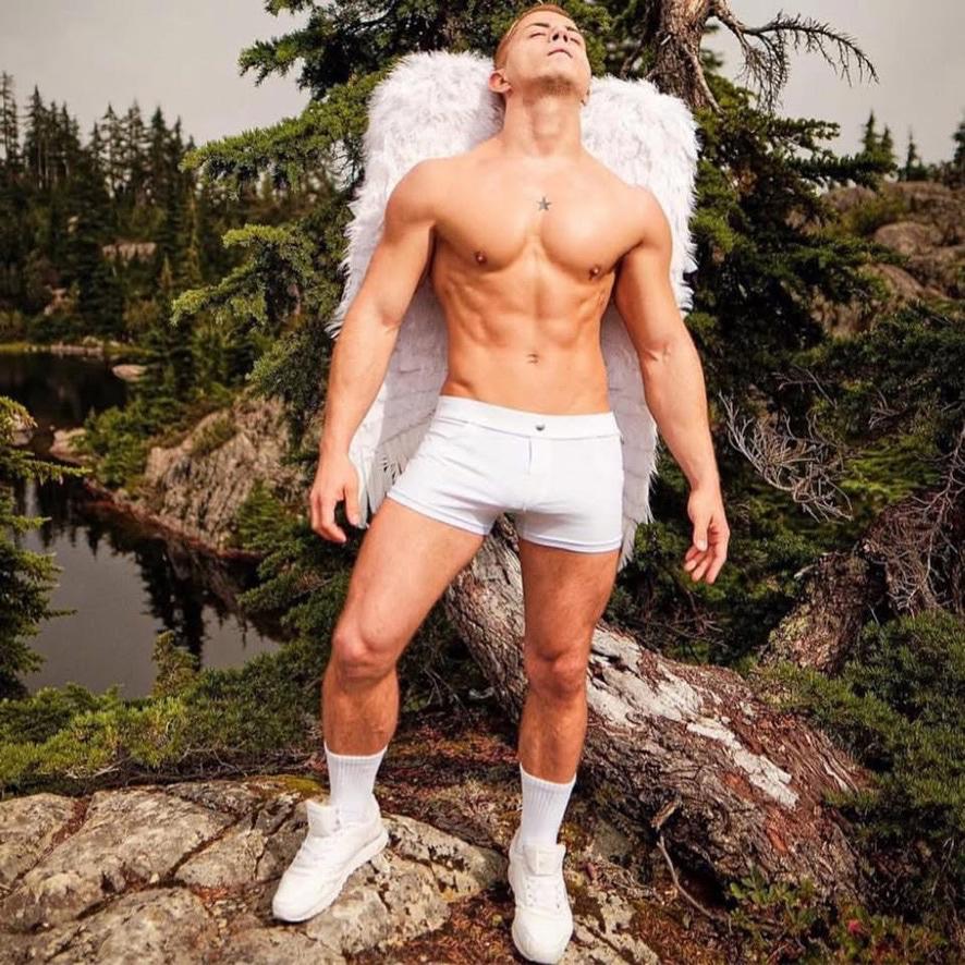 Damon posing as a muscle model in white underwear with large white feather wings in a mountain photoshoot near Vancouver, Canada